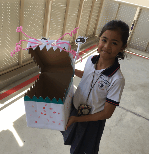 A smiling girl in school uniform holds a cardboard box decorated as a monster with jagged teeth and pink pipe cleaners.